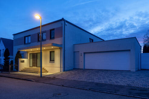 Deutschland, Bayern, Straßenlaterne leuchtet vor modernem Vorstadthaus in der Abenddämmerung - MAMF02740