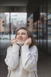 Happy woman holding turtleneck collar on footpath - IHF01333