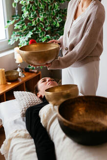 Ein Therapeut gibt einer Frau, die auf einer bequemen Massageliege im Spa liegt, eine Klangtherapie mit tibetischen Klangschalen - ADSF43512