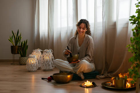 Ganzkörperaufnahme einer positiven jungen Frau mit lockigem dunklem Haar in Freizeitkleidung, die mit gekreuzten Beinen auf einem Hocker auf dem Boden sitzt und während der Meditation in einer Wohnung eine tibetische Klangschale spielt - ADSF43514