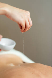 Crop anonymous therapist pouring warm oil on naked back of relaxed female client lying on table during massage session in light salon - ADSF43535