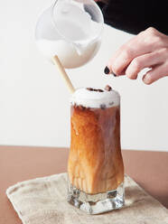 Unrecognizable person adding milk into glass of delicious coffee with whipped cream and straw on napkin in studio - ADSF43547