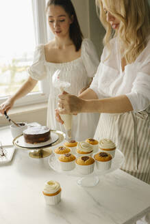 Frau mit Freundin bei der Zubereitung von Kuchen und der Dekoration von Törtchen zu Hause - VIVF00458