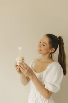 Glückliche junge Frau hält Geburtstagstörtchen mit brennender Kerze - VIVF00464