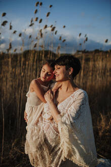 Lächelnde schwangere Frau mit Tochter auf einem Feld bei Sonnenuntergang - GMLF01378