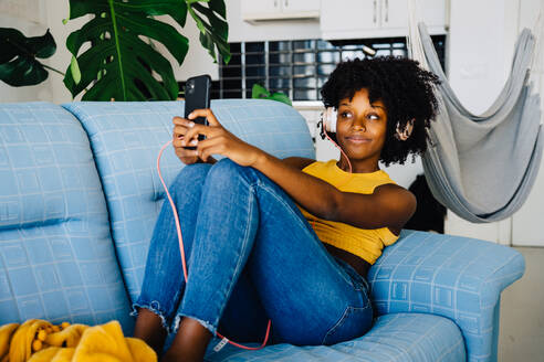 Eine entspannte junge Afroamerikanerin mit Afrofrisur macht ein Selfie, während sie Musik hört und sich in Freizeitkleidung auf dem Sofa räkelt - ADSF43620