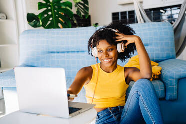 Lächelnde junge afroamerikanische Freiberuflerin in Freizeitkleidung, die auf dem Boden sitzt und auf den Bildschirm schaut, während sie mit Kopfhörern auf dem Laptop surft - ADSF43623