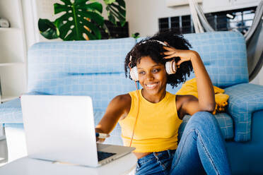 Smiling young African American female freelancer in casual outfit sitting on floor and looking at screen while browsing laptop in headphones during work - ADSF43623