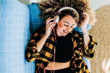 Top view of young female in casual clothes lying on blue couch while listening to music on headphones - ADSF43627