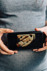 Abgeschnittene, nicht erkennbare, ruhige schwangere Frau in grauem Strickkleid, die vor einer weißen Wand steht und ein Bild des Fötus auf einem Smartphone in der Nähe des Bauches zeigt - ADSF43629