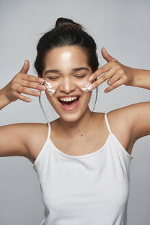 Positive junge Frau in weißem Oberteil mit geschlossenen Augen, die Feuchtigkeitscreme auf ihr Gesicht aufträgt - ADSF43640