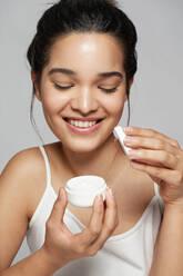 Erfreute junge Frau in weißem Oberteil lächelt und schaut nach unten, während sie eine Flasche mit einem Kosmetikprodukt öffnet - ADSF43642