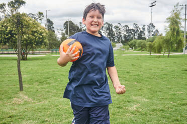 Cheerful preteen boy in sportswear with football smiling and celebrating victory while standing on grassy lawn - ADSF43646