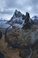 Luftaufnahme einer asphaltierten Straße zwischen verschneiten Bergen und blauem Meer unter bewölktem Himmel in Eystrahorn, Krossanesfjall, Island - ADSF43647