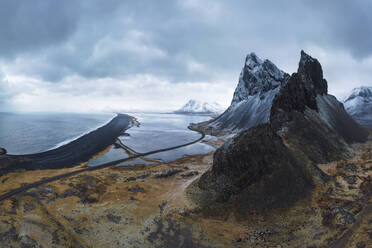 Luftaufnahme einer asphaltierten Straße zwischen verschneiten Bergen und blauem Meer unter bewölktem Himmel in Eystrahorn, Krossanesfjall, Island - ADSF43650