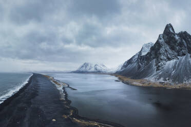 Drohnenansicht von oben auf die schneebedeckten Felsenberge in der Nähe des wogenden Meerwassers an der Südküste Islands gegen den bewölkten Himmel in Eystrahorn, Krossanesfjall, Island - ADSF43653