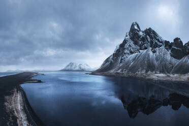 Drohnenansicht von oben auf die schneebedeckten Felsenberge in der Nähe des wogenden Meerwassers an der Südküste Islands gegen den bewölkten Himmel in Eystrahorn, Krossanesfjall, Island - ADSF43655