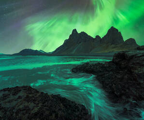 Erstaunliche Ansicht der grünen Aurora Borealis leuchtenden in den Nachthimmel über schneebedeckten Bergkamm mit schwarzem Sand Stockness Strand und Vestrahorn Berg im Hintergrund in Island - ADSF43660