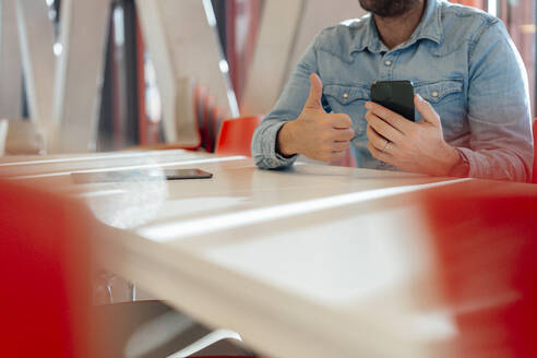 Geschäftsmann mit Smartphone und Daumen hoch am Tisch sitzend - JOSEF18151