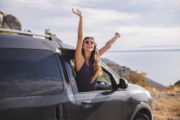 Cheerful young woman with arms raised leaning out of car window - PCLF00310