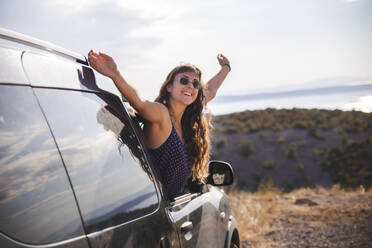 Happy young woman with arms raised leaning out of car window - PCLF00311