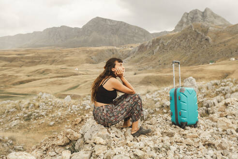 Verwirrte Frau mit Koffer auf einem Felsen sitzend - PCLF00338