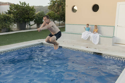 Mann springt in Schwimmbad mit Sohn und Tochter sitzen im Hintergrund - VIVF00473