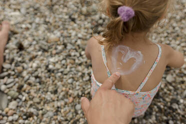 Man drawing heart shape with suntan lotion on daughter at beach - VIVF00524