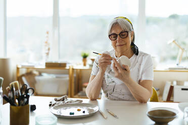 Lächelnde ältere Frau, die im Atelier eine Tasse bemalt - EBSF03076