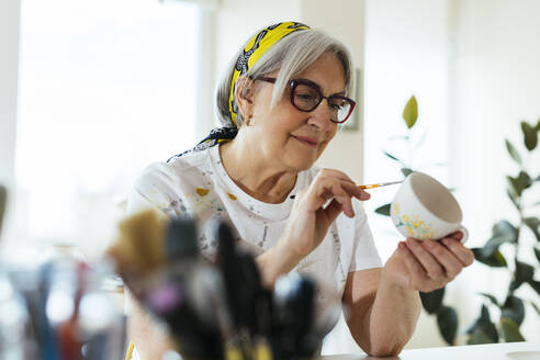 Lächelnde Frau mit Brille beim Bemalen einer Keramiktasse in einer Werkstatt - EBSF03078
