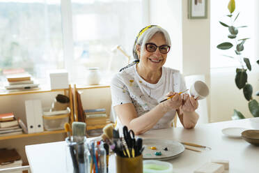 Glückliche ältere Frau mit Pinsel und Keramiktasse in der Werkstatt sitzend - EBSF03079