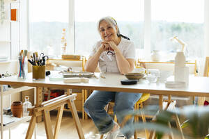 Glückliche ältere Frau sitzt mit Hand am Kinn am Tisch in einer Werkstatt - EBSF03080