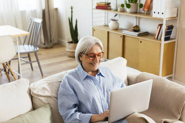 Lächelnde ältere Frau mit Laptop im Wohnzimmer sitzend - EBSF03114