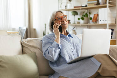 Lächelnde Frau, die mit ihrem Laptop im Wohnzimmer sitzt und mit ihrem Smartphone spricht - EBSF03115