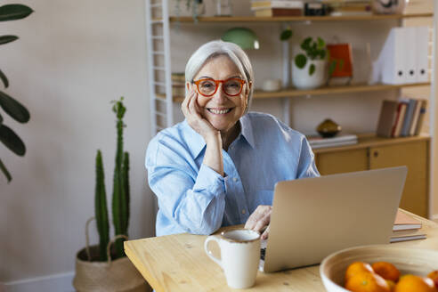 Happy senior woman sitting with hand on chin at home - EBSF03119