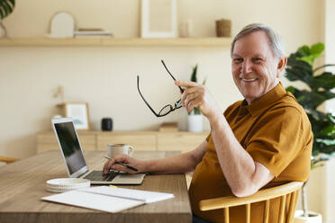 Smiling senior man sitting with laptop on table at home - EBSF03123