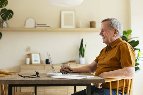 Lächelnder älterer Mann sitzt mit Laptop auf dem Tisch im Büro - EBSF03124