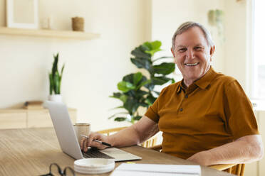 Glücklicher älterer Mann sitzt mit Laptop auf dem Tisch zu Hause - EBSF03125
