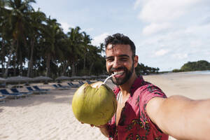 Ein fröhlicher Mann nippt an Kokosnusswasser und macht ein Selfie, während er die schöne Strandlandschaft genießt - PNAF05175