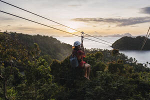 Woman riding on zip line over tree at sunset - PNAF05194