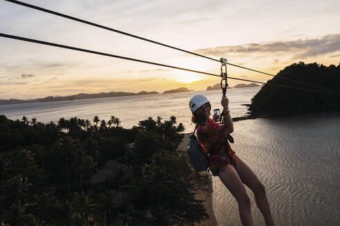 Happy woman enjoying on zip line over beach at sunset - PNAF05195