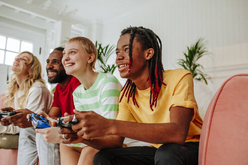 Lächelnder junger Mann mit Dreadlocks, der bei Freunden zu Hause ein Videospiel spielt - MDOF00939