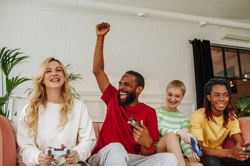 Aufgeregter junger Mann mit erhobener Hand, der zu Hause mit Freunden ein Videospiel spielt - MDOF00944