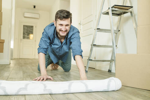 Happy young man unrolling carpet at new home - OSF01492