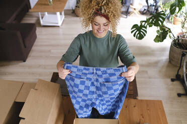 Happy young woman holding checked jeans at desk - ASGF03465