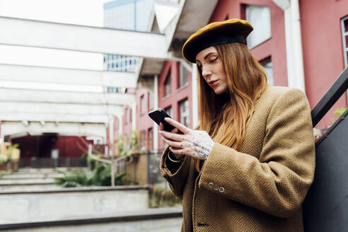 Rothaarige Frau, die mit einem Mobiltelefon vor einem Gebäude Textnachrichten verschickt - MEUF09073