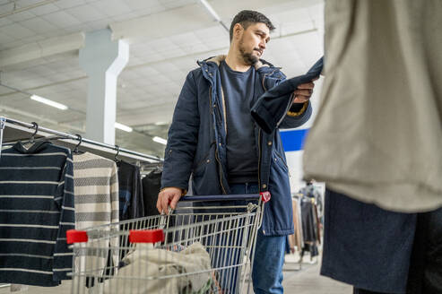 Mann mit Einkaufswagen betrachtet Kleidung in einem Geschäft - ANAF01169