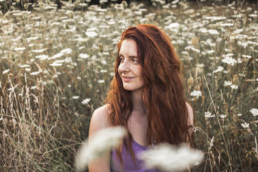Lächelnde rothaarige junge Frau inmitten von weißen Blumen auf einem Feld - PCLF00343
