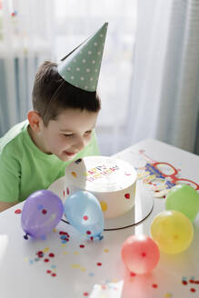 Lächelnder Junge betrachtet Geburtstagskuchen inmitten von Luftballons und Konfetti - ONAF00458