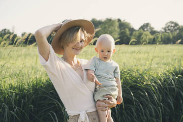 Happy mother wearing hat carrying son in nature on sunny day - NDEF00444